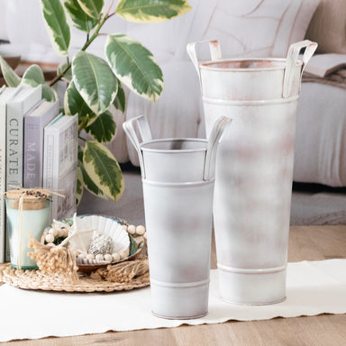 Two metallic vases on a table with decorative items and a plant in the background.